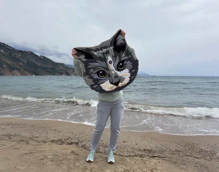 Artist - Kseniia Derr holding handcrafted detailed gray cat face rug against serene seaside background