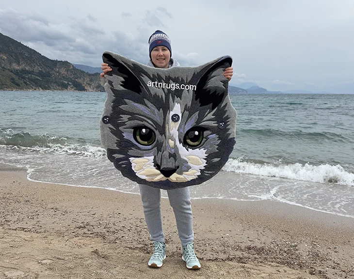 Artisan presenting handcrafted detailed gray cat portrait rug on coastal beach