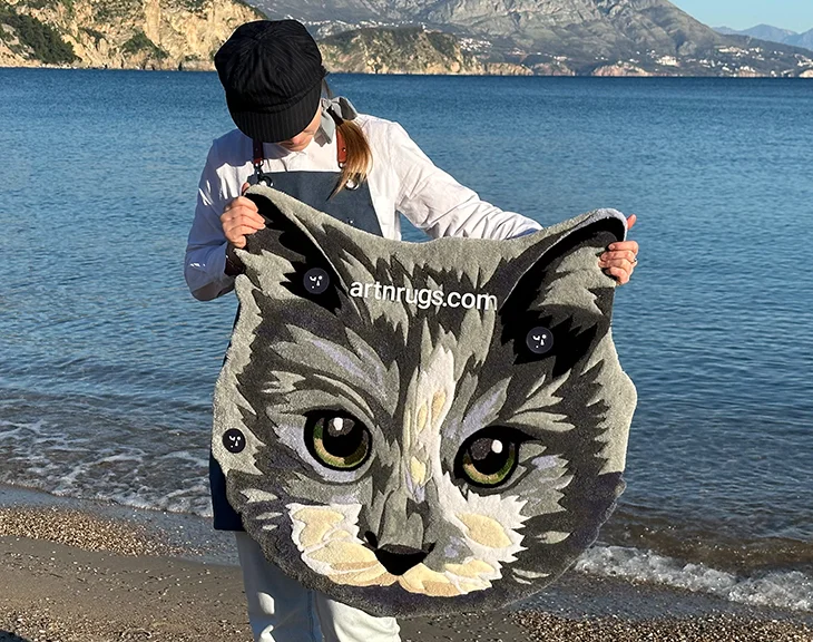 Artisan showcasing handcrafted detailed gray cat face rug in natural sunlight by the sea