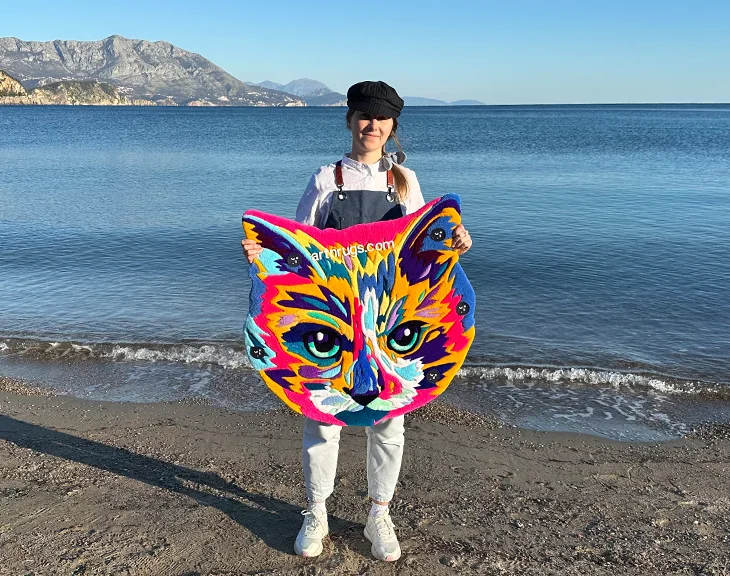 Woman holding vibrant multicolor cat face rug against clear seaside backdrop