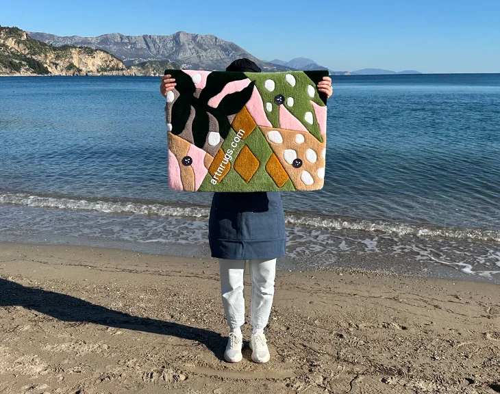 Eine Person steht am Sandstrand und hält einen modernen Tropenblatt-Teppich mit Grün-, Rosa-, Oliv- und Senfgelb-Formen und weißen ovalen Flecken hoch, im Hintergrund Berge und ruhiges, blaues Meer